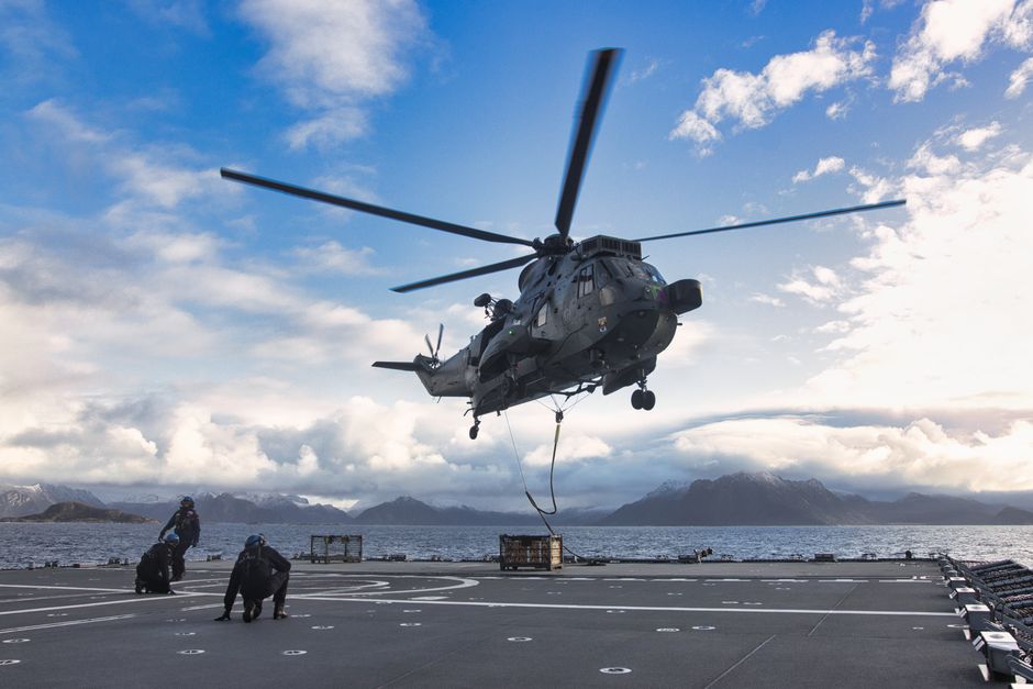 A military helicopter lands on a ship with mountains in the backdrop.