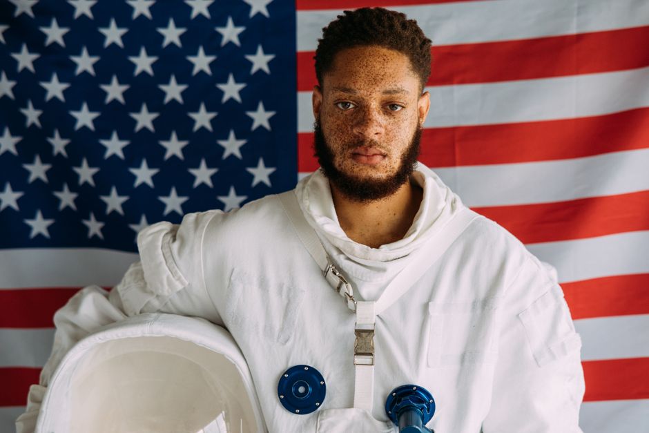 Man in astronaut suit standing against an American flag backdrop, showcasing a space-inspired theme.