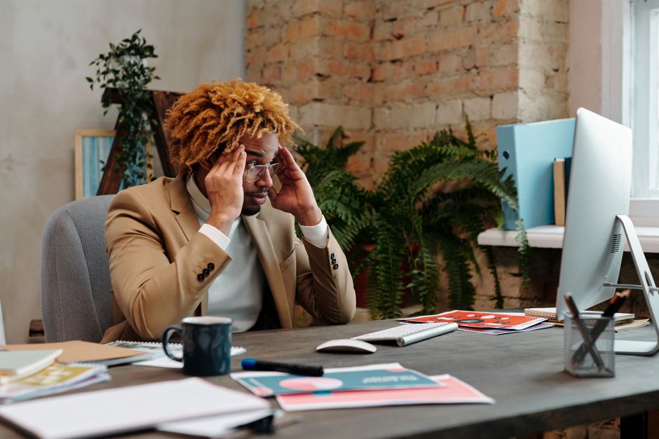 A businessman in stress at his office desk, overwhelmed with work documents and computer monitor.